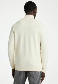 Man in een crèmekleurige hoge nektrui en vervaagde zwarte jeans, staande met de rug naar de camera tegen een effen witte achtergrond.