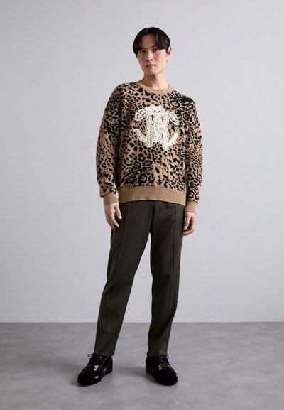 Pull en imprimé léopard beige avec des détails noirs et un logo blanc, associé à un pantalon vert foncé et des chaussures noires. Texture de tissu lisse.