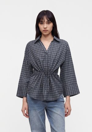 Jeune femme portant une chemise à carreaux grise boutonnée, cintrée à la taille avec des manches larges, associée à un jean bleu clair, debout devant un fond uni.