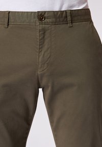 Pantaloni khaki realizzati in tessuto leggero con una texture liscia. Presentano una chiusura frontale con bottone e una vestibilità su misura. Cuciture doppie.