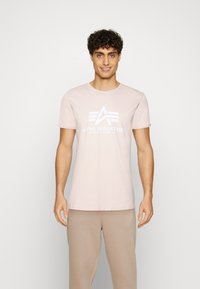 Gaiši rozā kokvilnas T-kreklā, ar baltu "ALPHA INDUSTRIES" grafiku un logo priekšpusē. Īsas piedurknes un apaļa kakla izgriezuma dizains.