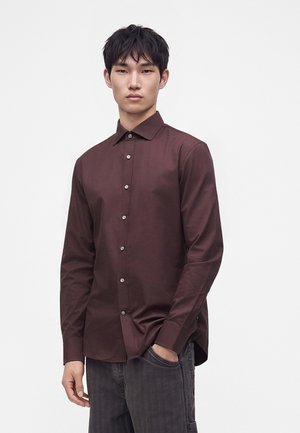 Young man with short black hair wearing a long-sleeve dark brown button-up shirt and dark gray pants with one hand in pocket.
