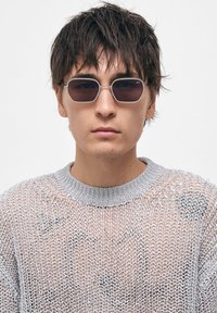 Aurinkolasit, joissa on tyylikkään hopeinen metallikehys, soikeat tummat linssit ja huomaamaton brändäys, yhdistettynä löysään hopeaan verkkoneulepaitaan.