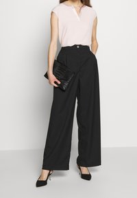 Helles rosa kurzärmliges Blusenshirt mit Schalkragen, kombiniert mit hochtaillierten schwarzen, weit geschnittenen Hosen und einer strukturierten schwarzen Clutch. Schwarze High Heels.