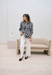Femme debout à côté d'un canapé beige, portant un chemisier à motifs noir et blanc, un pantalon blanc et des chaussures foncées dans une pièce minimaliste avec une statue de buste.