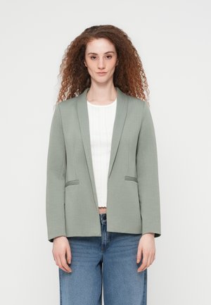 Vrouw met krullend haar die een saliegroene blazer draagt over een wit T-shirt en blauwe jeans, staand tegen een effen witte achtergrond.