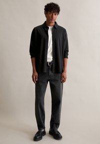 Čierna košeľa na gombíky cez biely tričko, v kombinácii s tmavými džínsami a čiernymi topánkami. Samostatná postava na neutrálnom pozadí.