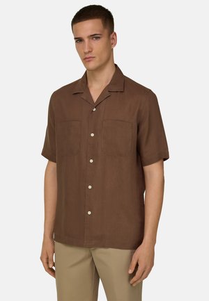 Junger Mann trägt ein braunes Kurzarmhemd mit zwei Brusttaschen und beigen Hosen vor einem einfarbigen Hintergrund.