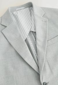 Blazer gris claro con una tela texturizada, presenta un forro interno a rayas, solapas con muescas y un solo bolsillo en el pecho.