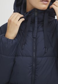 Veste matelassée marine avec une capuche, dotée d'une fermeture éclair centrale, de cordons de serrage réglables et d'un design matelassé pour l'isolation et la texture.