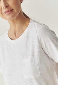 T-shirt bianca in cotone con colletto rotondo e una piccola tasca sul lato sinistro del petto; il tessuto ha una texture sottile e una finitura morbida.