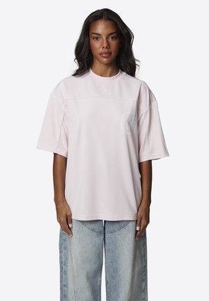 Vrouw met lang donker haar, gekleed in een loszittend lichtroze gaas shirt met korte mouwen en lichtblauwe jeans met wijde pijpen, staand en naar voren gericht.