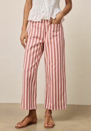 Frau trägt hoch taillierte, weit geschnittene Hose mit vertikalen rosa-weißen Streifen, kombiniert mit einem weißen, gerafften Oberteil und braunen, geflochtenen Sandalen.