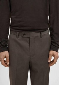 Pantalones marrones con estampado y corte recto, con cierre de botón y trabillas para cinturón. La textura parece suave con un diseño sutil de cuadrícula.