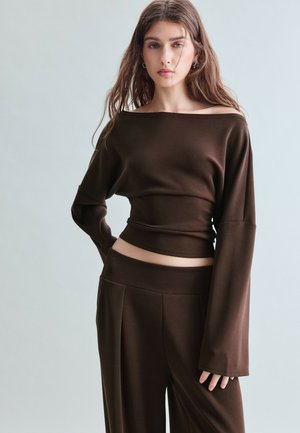 Ung kvinde med langt brunt hår iført en mørkebrun off-shoulder top og matchende vidde bukser, stående med den ene hånd på hoften.