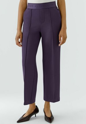 Vrouw draagt donkerpaarse, rechte, hoog getailleerde broek met voornaad en zwarte puntige hakken tegen een effen achtergrond.