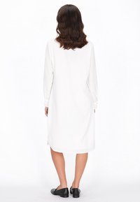 Witte lange mouw jurk met een rechte silhouette, voorzien van zijsplitten en een soepele textuur, in combinatie met zwarte platte schoenen.