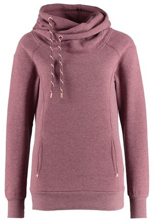 Person iført en mauve hættetrøje, blå jeans, sorte og hvide sneakers og bærende på en sort skuldertaske.