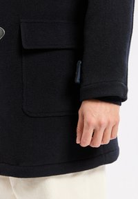 Main reposant à côté d'un manteau en laine noir avec une grande poche avant et des boutons bleus, porté sur un pantalon couleur crème.