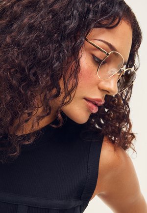 Mujer con cabello rizado oscuro que lleva grandes gafas redondas doradas y una camiseta negra sin mangas, mirando hacia abajo a la derecha.