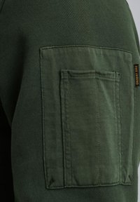 Groene sweatshirtstof met een gestructureerde zak op de linkermouw. Bevat een klein zwart label met "PME Legend" op de zak.