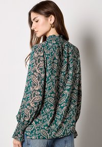 Langarmbluse mit grünem Grund, die ein Paisleymuster in Creme und Taupe aufweist. Geraffter Hals- und Handgelenkbereich aus leichtem, strukturiertem Stoff.