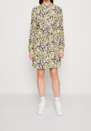 Blumenkleid mit gelben und lila Blumen auf schwarzem Hintergrund, langen Ärmeln, geraffter Taille und knielangem Saum, kombiniert mit weißen Turnschuhen.