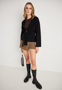 Čierny cardigan so šnúrkou v páse, skombinovaný s hnedým topom a mini sukňou s houndstooth vzorom. Doplňujú outfita čierne čižmy po kolená.
