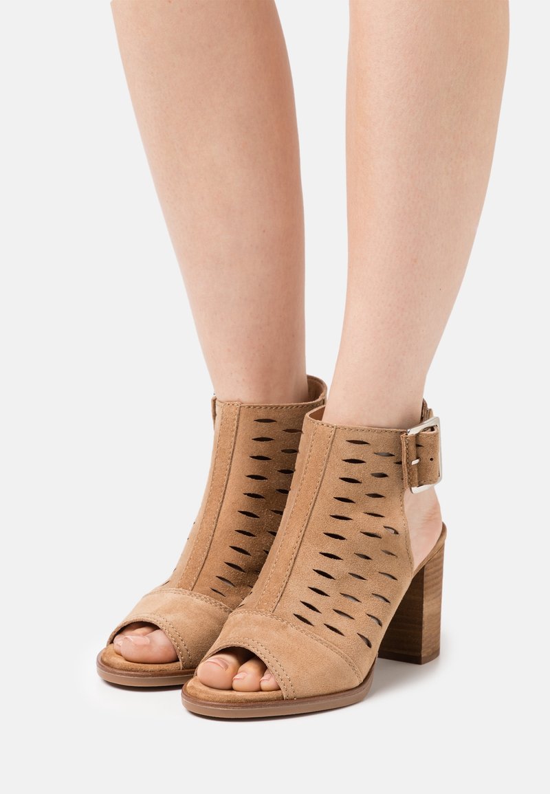 Bottines en daim beige à bout ouvert avec détails découpés, une bride latérale à boucle et des talons blocs empilés portés pieds nus.