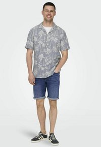 Camisa de botones de manga corta, gris claro, con un patrón de palmeras y veleros en blanco, combinada con pantalones cortos de mezclilla enrollados y zapatillas negras.