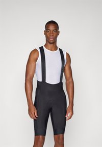 Schwarze Radsport-Bib-Shorts mit breiten Trägern, die ein weißes Mesh-Unterhemd tragen. Glatter Stoff mit gepolstertem Sitz und kontrastierenden elastischen Bändern am Saum.
