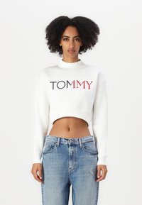 Fehér cropped pulóver magas nyakkal és tengerészkék, valamint piros "TOMMY" logóval. Világoskék, enyhén kopott farmernadrággal párosítva.