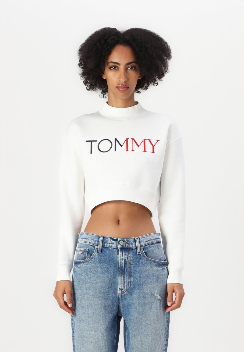 Fehér cropped pulóver magas nyakkal és tengerészkék, valamint piros "TOMMY" logóval. Világoskék, enyhén kopott farmernadrággal párosítva.