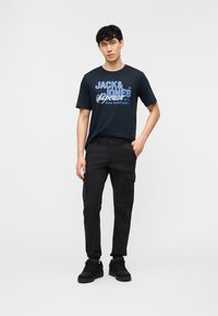 Giovane uomo in piedi che indossa pantaloni cargo neri, scarpe da ginnastica nere e una T-shirt nera con la scritta "Jack & Jones" in blu e bianco.