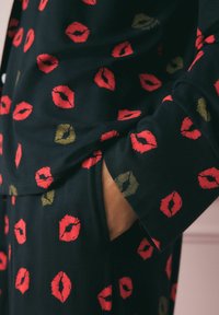 Mustaline kangas, millel on korduv punaste ja kuldsete suudluse jälgede muster. Disain on sujuv, vabam lõige, näidates kätt taskus.