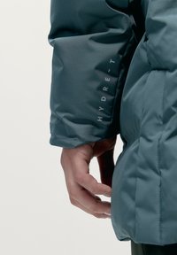 Giacca imbottita in verde scuro, con un esterno liscio e resistente all'acqua. Le caratteristiche includono un polsino piegato e un branding discreto sulla manica.