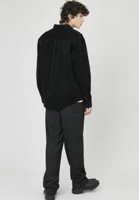 Chemise en velours côtelé noir à coupe décontractée, avec un détail de pli au dos, des manches longues et des poignets à bouton, associée à un pantalon noir ajusté et des chaussures épaisses.