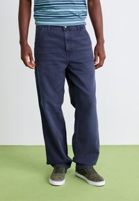 Man wearing loose dark blue pants, striped blue shirt, and gray sneakers standing on green floor against white wall.