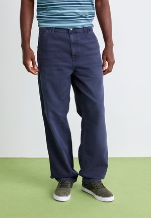 Man wearing loose dark blue pants, striped blue shirt, and gray sneakers standing on green floor against white wall.