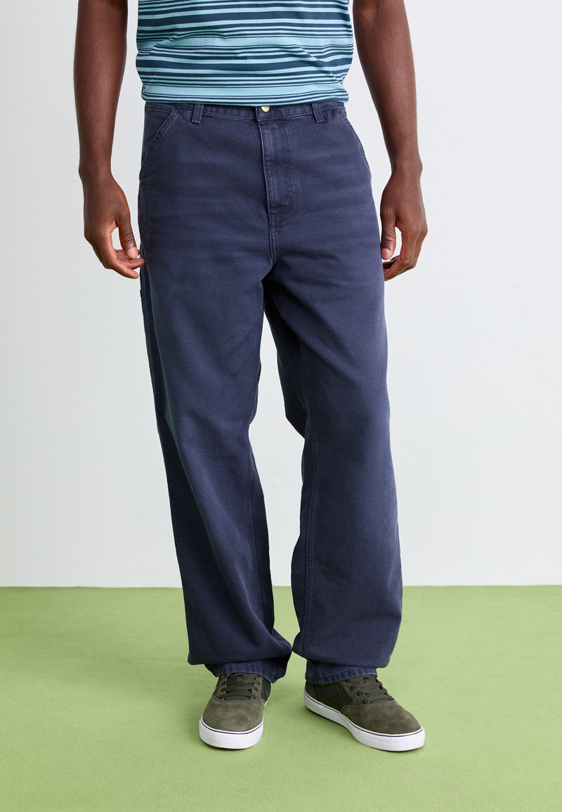 Man wearing loose dark blue pants, striped blue shirt, and gray sneakers standing on green floor against white wall.