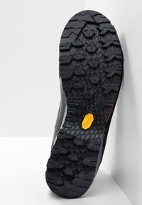 Svart gummi såle med dype spor for grep og godt feste, med en synlig gul logo detalj. Robust og teksturert design, skrå vinkel.