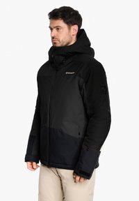 Schwarze isolierte Jacke mit Kapuze, ausgestattet mit einem kontrastierenden dunkelblauen unteren Bereich, mit Reißverschlusstaschen und einem Logo auf der Brust und dem Ärmel.