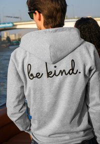 Felpa grigia con cappuccio, decorata con il testo in corsivo nero "be kind." sul retro. Materiale morbido, design casual, con maniche lunghe e vestibilità tranquilla.
