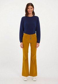 Pull marin et pantalon en velours côtelé moutarde évasé avec un devant à cinq boutons. Le mannequin porte des baskets blanches. Le fond uni met en valeur la tenue.
