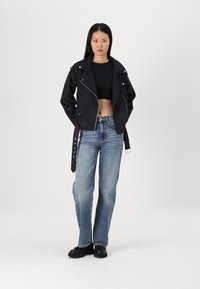 Black cropped jacket with silver zipper, collar, and belted waist, paired with light blue straight-leg jeans and black platform shoes.