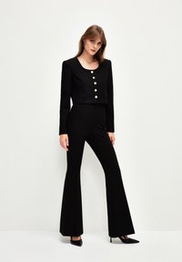 Zwarte cropped blazer met gouden knopen, gecombineerd met uitlopende zwarte broeken en zwarte hoge hakken; een zachte textuur overal.
