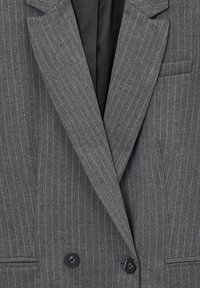 Blazer gris à rayures, avec des boutons noirs notables, des revers structurés et une texture lisse. Il dispose d'une poche poitrine et d'une coupe ajustée.