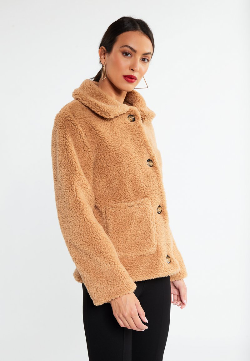 Beige Kunstpelzjacke mit einem Kragen, vier großen schwarzen Knöpfen und einer Vordertasche. Weicher Stoff und lässige Passform.