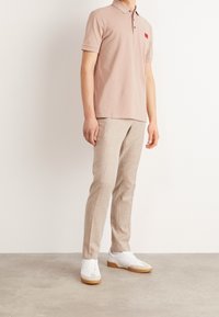 Jasnoróżowa koszulka polo z krótkim kołnierzykiem, z małym czerwonym logo. Beżowe spodnie o teksturowanej fakturze i białe sneakersy z gumową podeszwą.