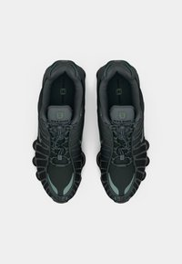 Par nogavica tamnozelene i crne Nike Shox atletske cipele s vezicama, prikazane odozgo na bijeloj pozadini.